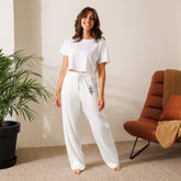 woman wearing white silky relaxed fit women’s pajama pants with decorative side seam piping and white cropped t-shirt indoors next to plant and chair