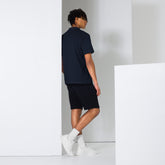 Man wearing navy blue men polo shirt with black shorts and white sneakers standing against white wall