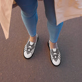 Women’s slip-on canvas shoes with black geometric patterns, worn with blue jeans on asphalt