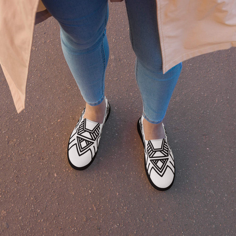 Women’s slip-on canvas shoes with black geometric patterns, worn with blue jeans on asphalt