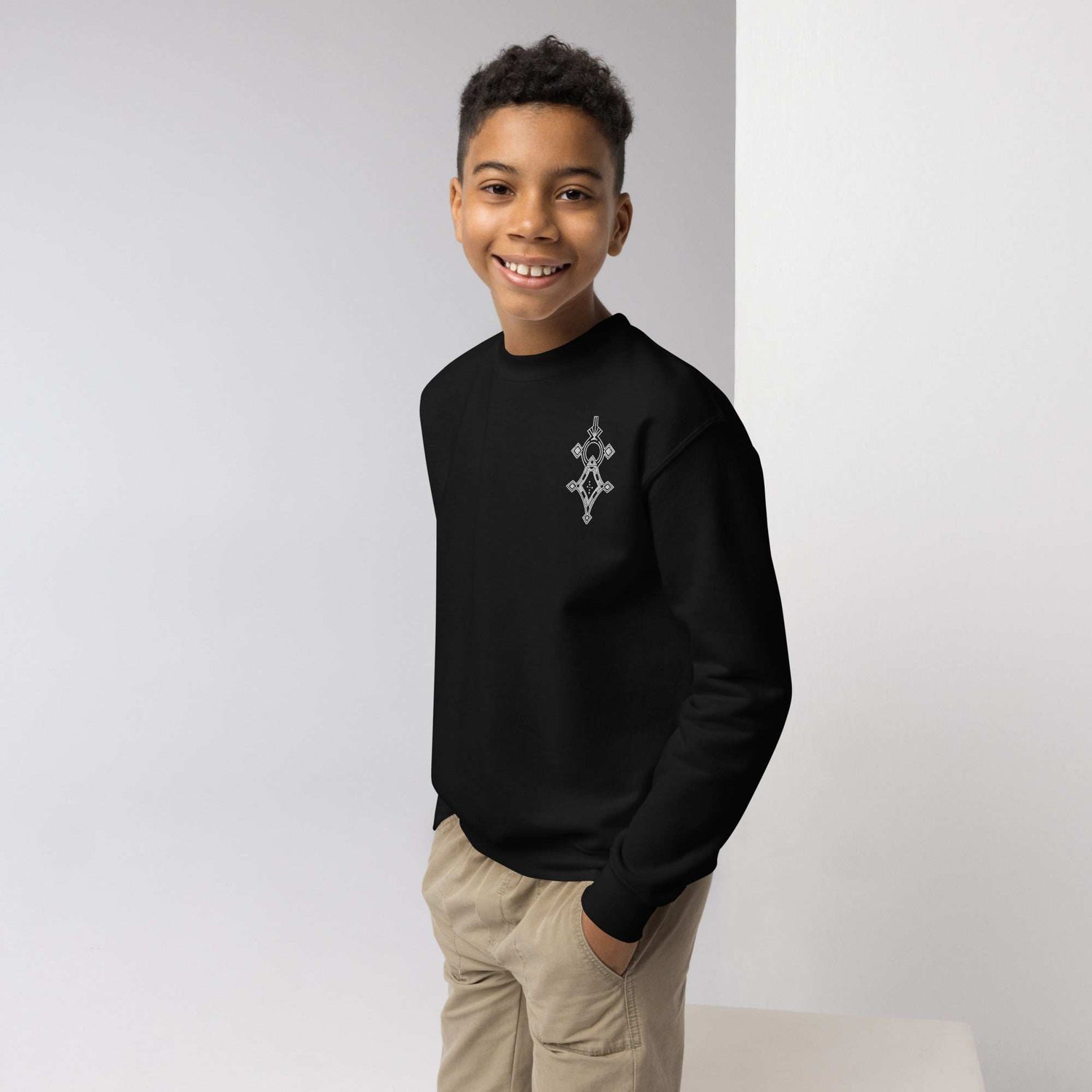 Smiling boy wearing black youth crewneck sweatshirt with geometric design, hands in khaki pants pockets, standing against gray and white background