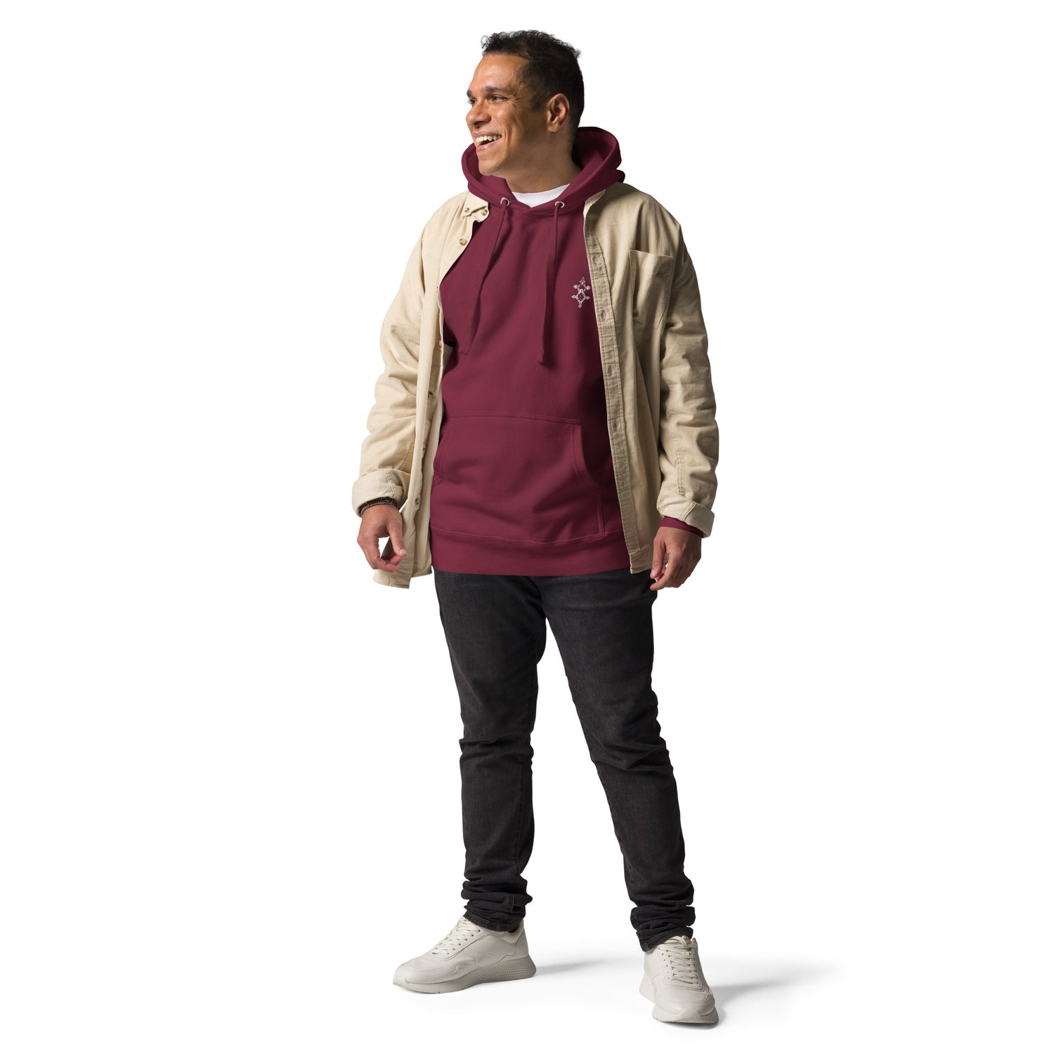 Man wearing a maroon men hoodie with front pouch pocket, beige jacket, black pants, and white sneakers standing against white background
