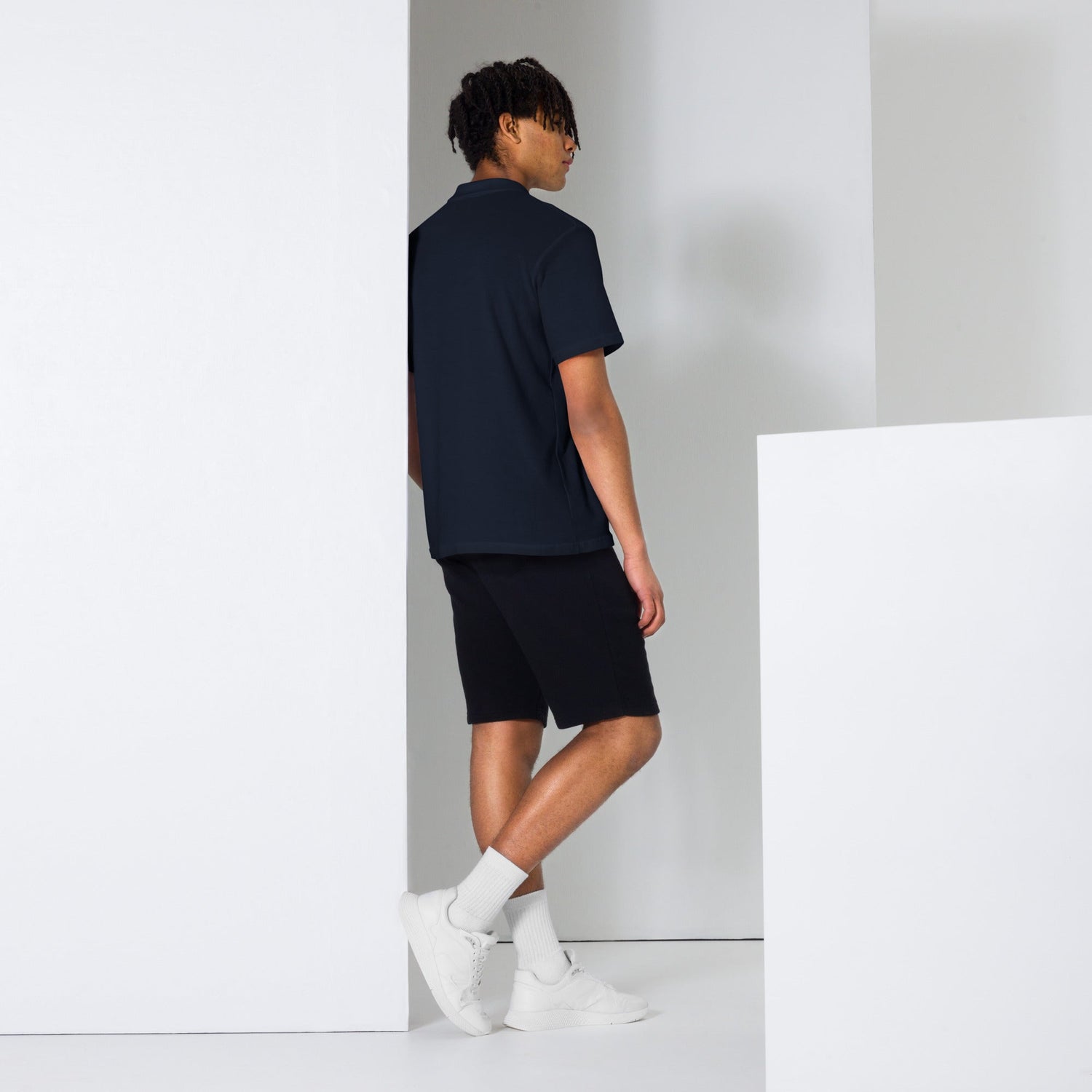 Man wearing navy blue men polo shirt with black shorts and white sneakers standing against white wall