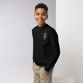 Smiling boy wearing black youth crewneck sweatshirt with geometric design, hands in khaki pants pockets, standing against gray and white background